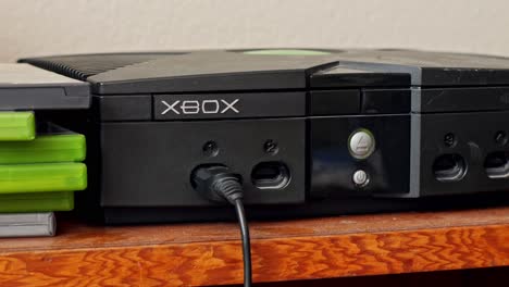 Conectar-El-Cable-Del-Controlador-E-Insertar-El-Disco-Del-Juego-En-Una-Consola-De-Videojuegos-Microsoft-Xbox-2001-Vintage-Sobre-Una-Mesa-De-Madera-Con-Estuches-De-Juegos-Xbox-Al-Lado