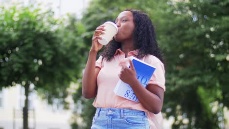 Student-Girl-Reading-Book-and-Drinking-Coffee.education,-school-and-people-concept-happy-smiling-african-american-student-girl-reading-book-and-drinking-takeaway-coffee-in-city