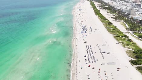 Drone-descends-from-high-altitude-to-close-to-the-shoreline-at-South-Beach,-capturing-people-in-the-water-and-waves