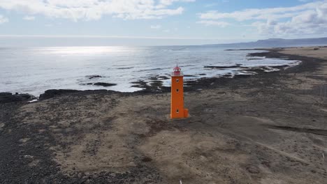 Der-Leuchtturm,-Der-Hoch-Auf-Einem-Schwarzen-Sandstrand-Steht,-Ist-Ein-Markantes-Merkmal-In-Diesem-Luftbildvideo-Der-Isländischen-Küste-Mit-Ihrer-Felsigen-Landschaft-Und-Den-Bergen-In-Der-Ferne