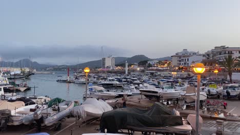 Yachthafen-Auf-Mallorca-In-Der-Abenddämmerung-Mit-Angedockten-Booten-Und-Sanft-Leuchtenden-Lichtern-Der-Stadt-Im-Hintergrund