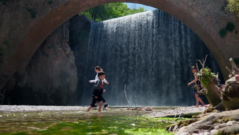 Junge-Leute-Spielen-Und-Genießen-Das-Wasser-Vor-Einem-Großen-Wasserfall-Im-Zeitlupen-Hintergrund-Mit-Bogensteinbrücke