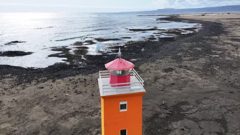 Luftaufnahmen-Zeigen-Einen-Leuchtend-Orangefarbenen-Leuchtturm-Mit-Rosa-Dach,-Der-Stolz-Auf-Einem-Schwarzen-Sandstrand-Entlang-Der-Zerklüfteten-Küste-Islands-Steht