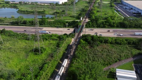 Train-crosses-tracks-above-bustling-highway-below-electricity-pylons,-aerial-dolly