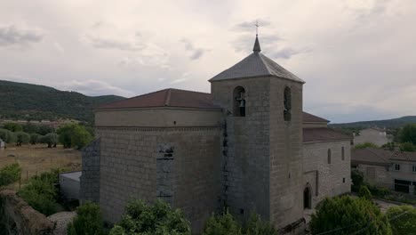 ascending-flight-with-a-drone-in-the-church-of-San-Bartolome-16th-century-in-the-back,-seeing-the-stone-back-with-its-buttresses,-the-bell-tower-and-discovering-the-fantastic-place-where-it-is-located