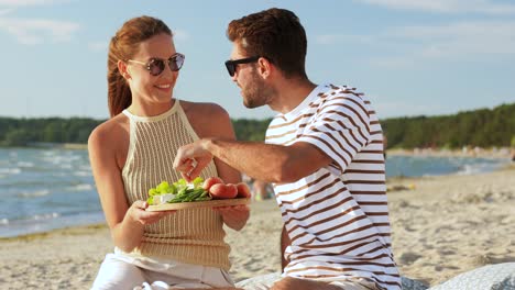 Happy-Couple-with-Food-Having-Picnic-on-Beach.leisure,-relationships-and-people-concept-happy-couple-with-food-eating-grapes-and-having-picnic-on-beach