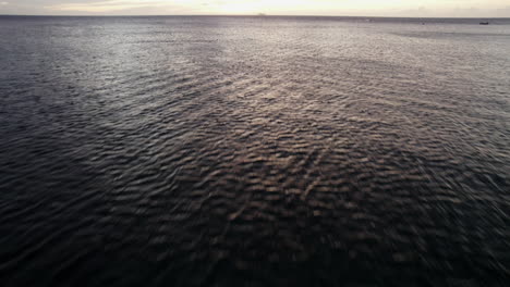 Aerial-view-flying-over-the-vast-oceanfront,-observing-water-ripples-stretching-towards-the-horizon-during-twilight-off-the-coast-of-Roatan,-Honduras