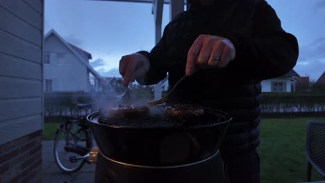 Hands-turning-a-burger-on-a-grill-with-flames,-fire,-and-smoke-in-a-garden-during-twilight