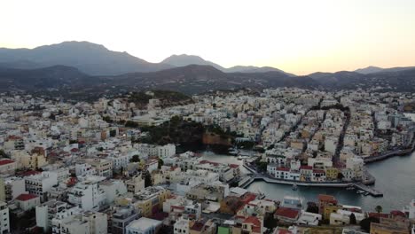 Aerial-Panoramic-over-Agios-Nikolaos-Town-and-Mountainous-Region-on-Background-at-Sunset,-Greece