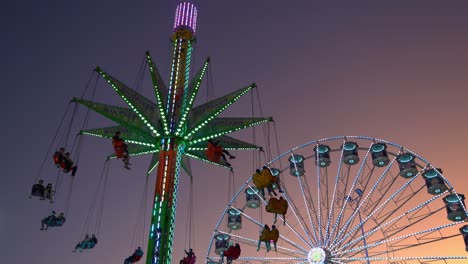 Illuminated-sky-flyer-festival-ride,-elevating-and-swinging-up-high-in-the-sky-against-sunset-dusk-sky-and-iconic-ferris-wheel-spinning