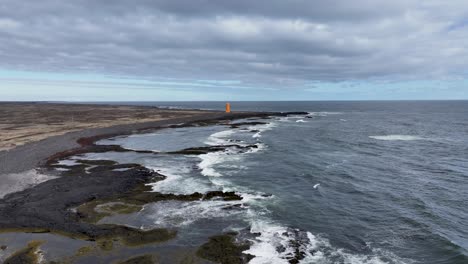 Ein-Luftbildvideo-Zeigt-Einen-Malerischen-Leuchtturm-An-Islands-Küste,-Dessen-Leuchtende-Orange--Und-Rosatöne-Sich-Vor-Der-Kulisse-Felsigen-Geländes-Und-Meereswellen-Abheben