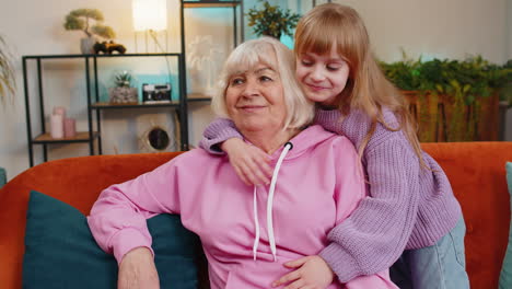 Cheerful-loving-small-girl-granddaughter-embracing-kissing-happy-grandmother-sitting-on-home-sofa