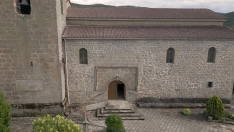 Drone-flight-visualizing-the-entrance-façade-to-the-16th-century-church-of-San-Bartolome,-its-late-Gothic-decoration,-the-entrance-porch-with-stairs,-the-bell-tower-and-a-background-of-mountains