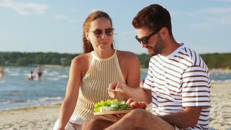 Happy-Couple-with-Food-Having-Picnic-on-Beach.leisure,-relationships-and-people-concept-happy-couple-with-food-eating-grapes-and-having-picnic-on-beach