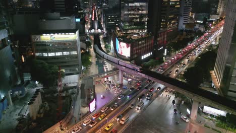 Drone-aerial-establishing-of-Mrt-skytrain-railway-at-night-in-Bangkok-Thailand-cityscape,-traffic-intersection-and-skyscrapers,-commercial-nighttime