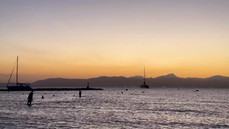 Menschen-Waten-Bei-Sonnenuntergang-Im-Seichten-Wasser,-Während-In-Der-Nähe-Auf-Mallorca-Boote-Vor-Anker-Liegen