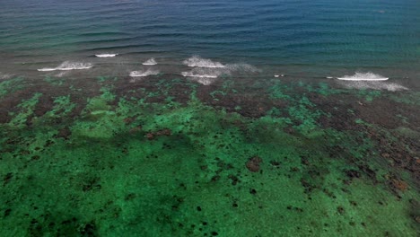 Drone-shot-of-deep-blue-water-crashing-into-shallower,-clear-waters-with-visible-coral-reefs