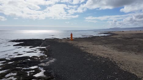 Drohnenaufnahmen-Fangen-Die-Schönheit-Eines-Farbenfrohen-Leuchtturms-An-Der-Küste-Islands-Ein,-Der-Auf-Einem-Schwarzen-Sandstrand-Inmitten-Einer-Dramatischen-Landschaft-Aus-Felsen-Und-Meer-Thront