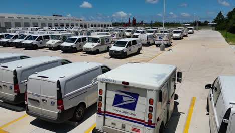 Toma-Aérea-De-Dron-Que-Se-Eleva-Lentamente-De-Los-Camiones-De-Reparto-De-USPS-Detrás-De-Una-Gran-Oficina-De-Correos-Desde-La-Esquina-Del-Estacionamiento,-Comenzando-Con-Un-Gran-Logotipo-De-USPS-En-La-Parte-Posterior-De-Un-LLV.