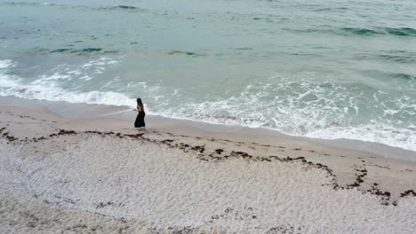 Schönes-Mädchen-Mit-Schwarzem-Kleid,-Das-Am-Meer-Entlang-Geht,-Mit-Wellen-In-Ihren-Füßen