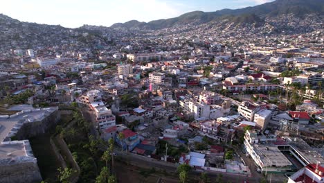 Backwards-scenic-flight-over-residential-area-in-Acapulco-with-crowded-cityscape,-mountains-and-coastal-road