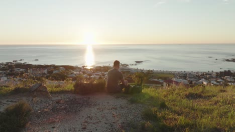 Drohne-Liegt-Langsam-Herum,-Männliche-Person-Sitzt-An-Einem-Hang-Am-Camps-Bay-Beach-In-Kapstadt,-Südafrika---Blick-Auf-Den-Strand-Und-Das-Meer-Bei-Sonnenuntergang