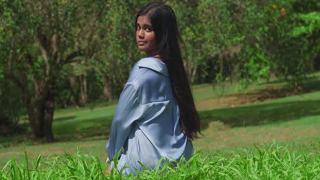 At-the-park,-a-young-East-Indian-girl-dressed-in-a-blue-shirt-on-a-sunny-day-kneel-in-the-grass