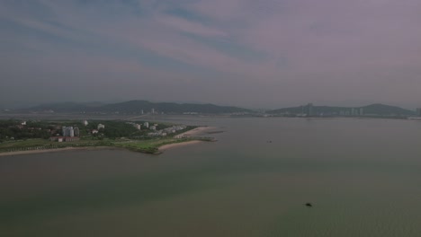 Drone-shot-flying-over-green-and-brown-waters-in-Ha-Long,-Vietnam,-towards-land-with-beaches,-a-bridge,-boat,-mountains,-and-tall-buildings-under-a-cloudy-sky