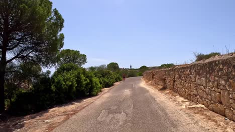 Person-walking-along-secluded-sloping-roadside,-point-of-view-journey-towards-Calamosca-Tower,-Cagliari