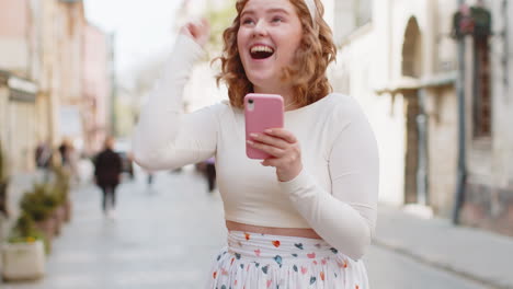 Young-woman-use-mobile-smartphone-celebrating-win-good-message-news-outdoors-in-urban-city-street