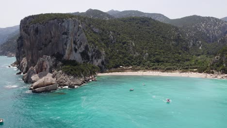 Barcos-Y-Gente-En-Una-Hermosa-Playa-Con-Grandes-Acantilados-En-Cerdeña,-Italia