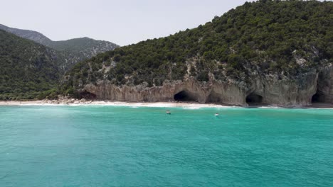 Aerial-Shot-Of-Boat-Drifting-In-Turquoise-Ocean,-People-Swimming-On-Sandy-Beach-In-Sardinia,-Italy