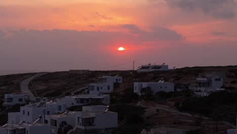 Drone-rises-above-white-homes-on-hillside-of-Donousa-Greece-as-fire-red-ball-of-sun-at-sunset-dips-to-hide-below-clouds-on-ocean