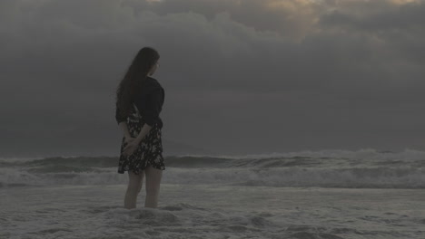 Pondering-young-female-wearing-glasses-looking-out-across-stormy-overcast-ocean-waves