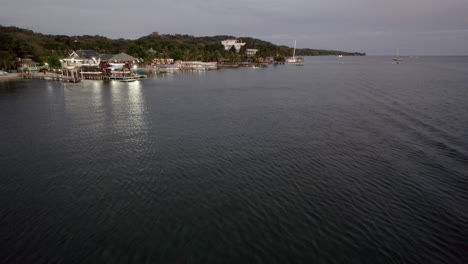 Fast-aerial-push-in-gliding-across-the-ocean,-moving-towards-docks-with-boats-on-the-beachline-during-dusk-in-Roatan,-Honduras