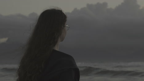 Mujer-Joven-Con-Gafas-Mirando-Las-Olas-Del-Océano-Nublado-Y-Tormentoso,-Soñando