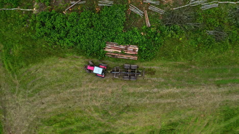 Drone-tops-down-field-tractor-and-piles-of-wood-neatly-cut-after-tree-deforestation,-shot-reveals-surroundings