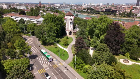 Ein-Grüner-Park-In-Basel-Mit-Einem-Historischen-Tor,-Einer-Straßenbahn-Und-Stadtbild-Im-Hintergrund,-Luftaufnahme