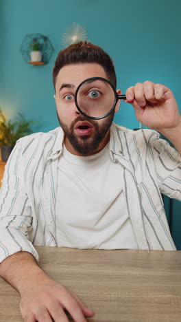 Freelancer-man-holds-magnifying-glass-near-face-looking-at-camera-with-big-zoomed-eye,-analyzing