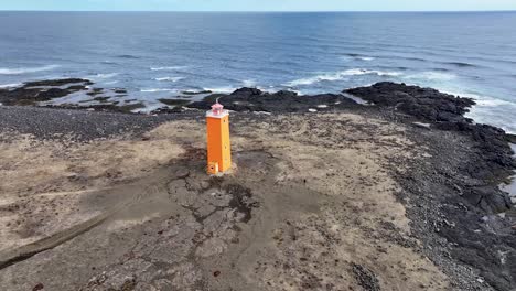 Luftaufnahme-Eines-Leuchtenden-Leuchtturms-An-Der-Küste-Islands,-Umgeben-Von-Einer-Zerklüfteten-Landschaft-Aus-Schwarzem-Sand,-Felsen-Und-Meereswellen,-Mit-Bergen-Im-Hintergrund