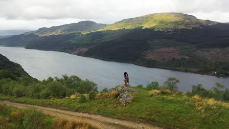 Aerial-pan-of-Scottish-landscape,-bagpiper-playing-on-a-mountain-above-loch-in-highland-vista