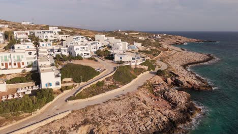 Un-Dron-Orbita-A-Lo-Largo-De-La-Costa-Rocosa-De-Donoussa,-Grecia,-Con-Casas-Elevándose-A-Lo-Largo-De-La-Ladera-Sobre-Un-Camino-Arenoso