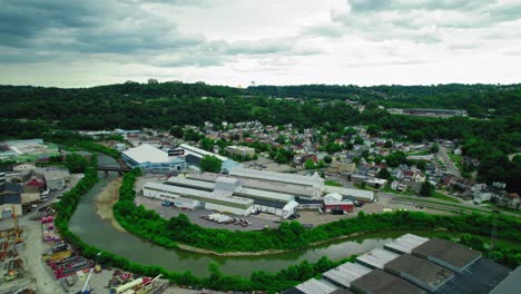 Aerial-view-of-the-industrial-area-in-Carnegie,-Pennsylvania,-featuring-warehouses,-residential-neighborhoods,-and-lush-green-hills-under-a-cloudy-sky