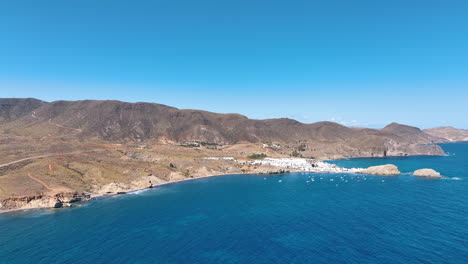 Flight-across-the-Mediterranean-sea-towards-the-moored-boats-and-white-walled-village-of-La-Isleta-del-Moro-with-the-arid-Monte-Ireder-in-the-background-on-a-perfect-hot-day-under-blue-skies