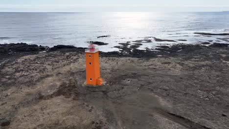 Ein-Luftbildvideo-Zeigt-Einen-Malerischen-Leuchtturm-An-Islands-Küste,-Dessen-Leuchtende-Orange--Und-Rosatöne-Sich-Vor-Der-Kulisse-Felsigen-Geländes-Und-Meereswellen-Abheben