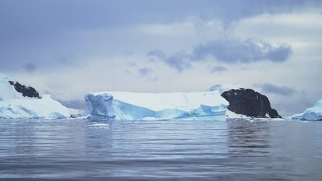 Pájaro-Volando-Con-Un-Iceberg-Azul-Al-Atardecer-En-El-Paisaje-De-La-Antártida,-Ave-Marina-En-Vuelo-En-Un-Paisaje-Invernal-Con-Mar-Oceánico-Y-Espectaculares-Nubes-Al-Amanecer,-Vida-Salvaje-Costera-En-La-Hermosa-Península-De-La-Antártida