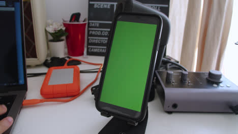 Beautiful-close-up-and-slow-motion-shot-of-a-mobile-phone-with-green-screen-for-animation-resting-on-a-pedestal-on-an-office-desk-with-a-white-table-and-a-computer-next-to-it