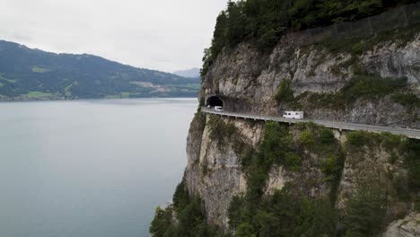 Camper-driving-along-a-cliffside-road-with-lake-view-in-Switzerland,-aerial-shot