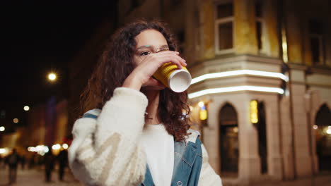 Happy-smiling-young-girl-enjoying-coffee-hot-drink-relaxing-taking-a-break-in-night-city-street