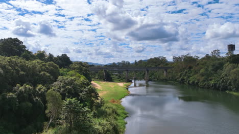 Aerial-lifts-up-from-riverside-lush-vegetation-and-above-the-grassed-banks-of-Mackay-region's-famous-Platypus-Beach-swimming-area-by-the-Pioneer-River-and-solitary-car-crossing-road-bridge-behind